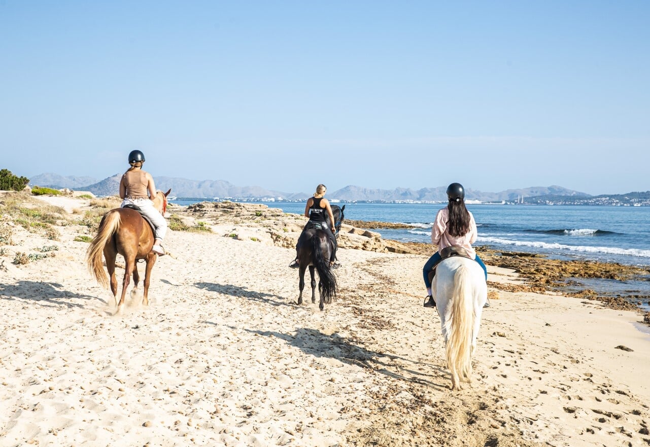 Drei Personen reiten am Strand am Meer auf einem schwarzen, braunen und weißen Pferd während eines Ausritts auf Mallorca